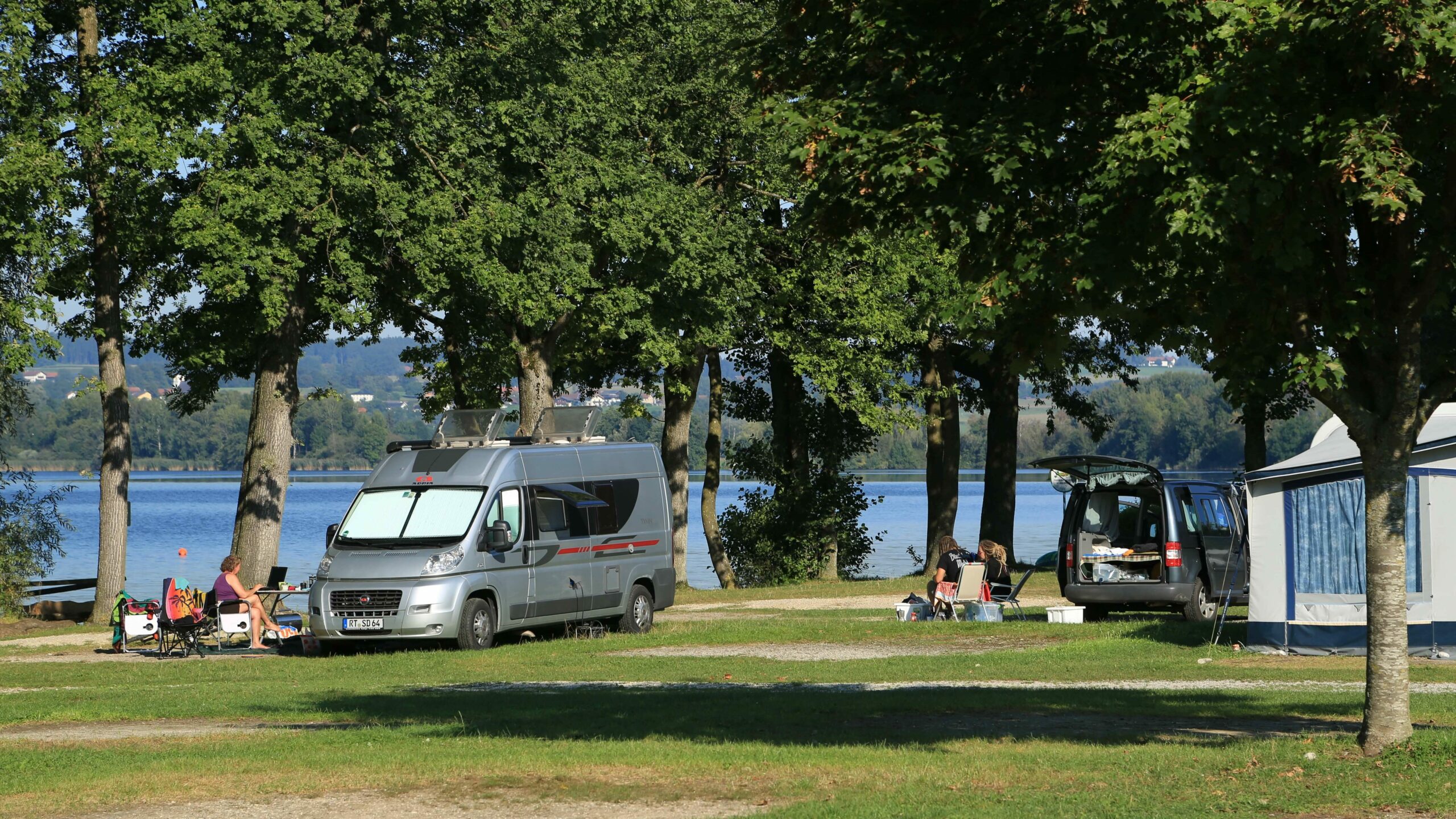 Mehrere Camper und Campingbusse auf der Campingwiese vor dem See.