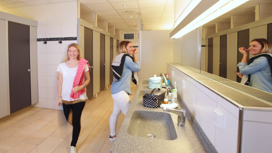 Zwei Frauen in den Sanitäranlagen des Campingplatzes. Links eine Reihe an Toiletten, rechts eine Reihe von Waschbecken mit einem langen, beleuchteten Spiegel.