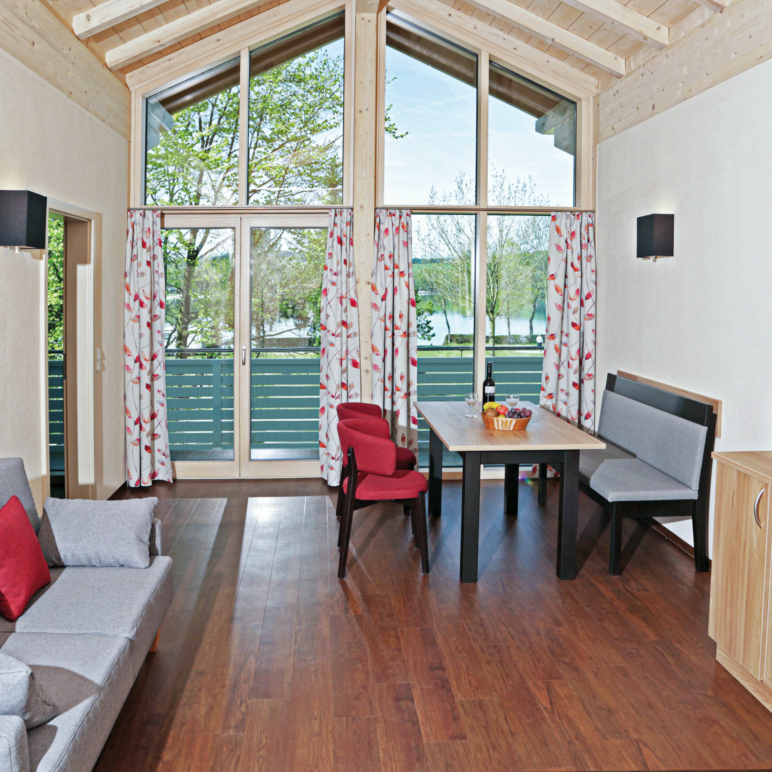 Das gemütlich eingerichtete Wohnzimmer einer Ferienwohnung. Hinten ist eine große Fensterfront, davor ein Esstisch mit 4 Stühlen und einer Bank, links ein Sofa, davor ein Fernseher.