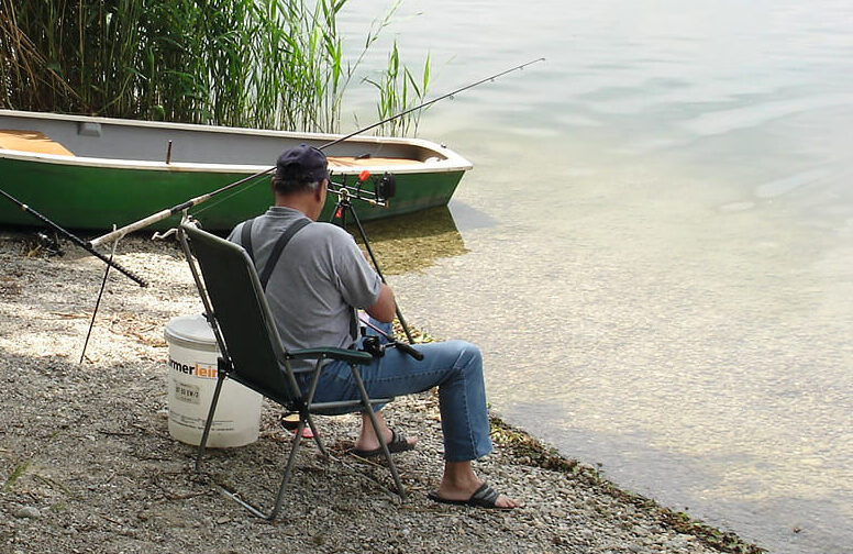 Ein Angler sitzt mit Angelausrüstung und einem kleinen Boot am Ufer des Sees