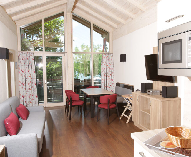 Das gemütlich eingerichtete Wohnzimmer einer Ferienwohnung. Hinten ist eine große Fensterfront, davor ein Esstisch mit 4 Stühlen und einer Bank, links ein Sofa, davor ein Fernseher und im Vordergrund ein Teil der Küche mit einer Mikrowelle.