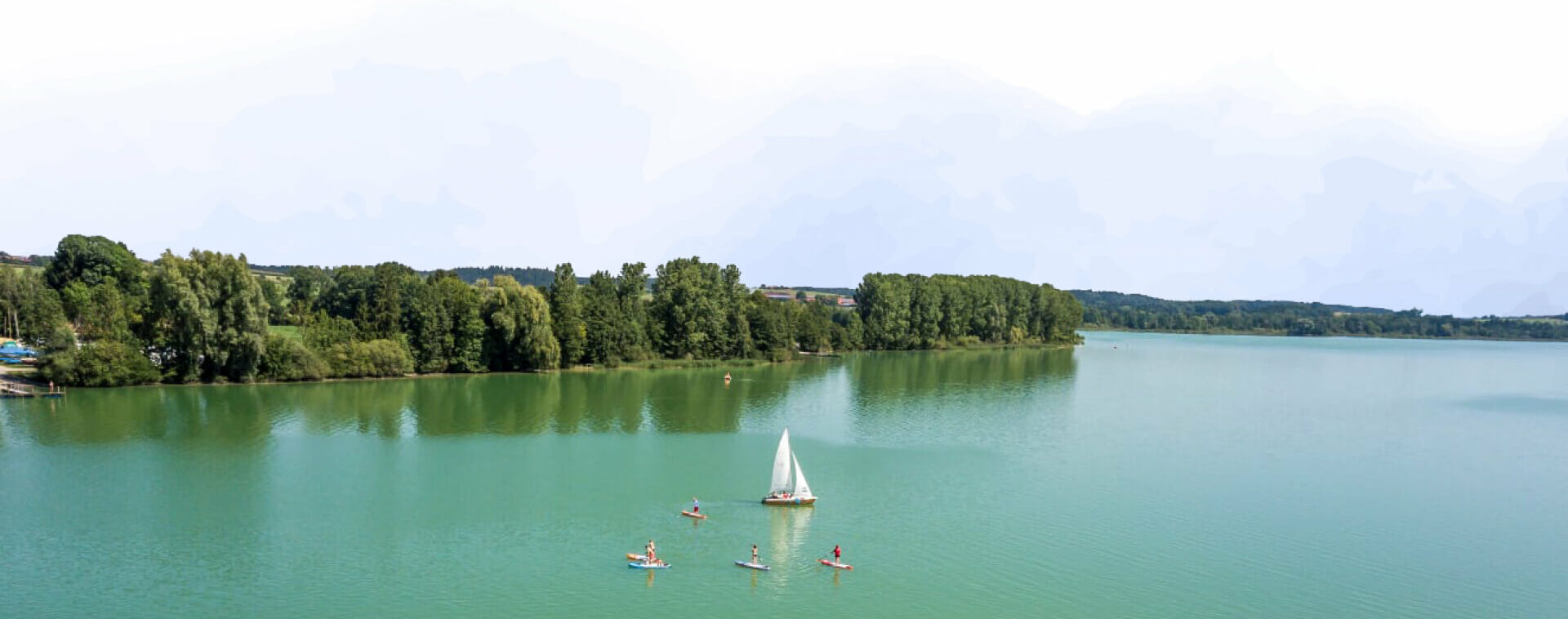 Der Waginger See von Oben, mittig treibt ein Segelboot umgeben von mehreren Stand-up-Paddlern
