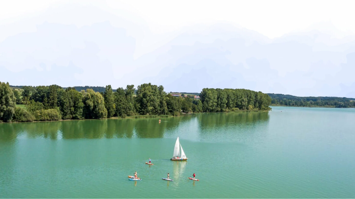 Der Waginger See von Oben, mittig treibt ein Segelboot umgeben von mehreren Stand-up-Paddlern