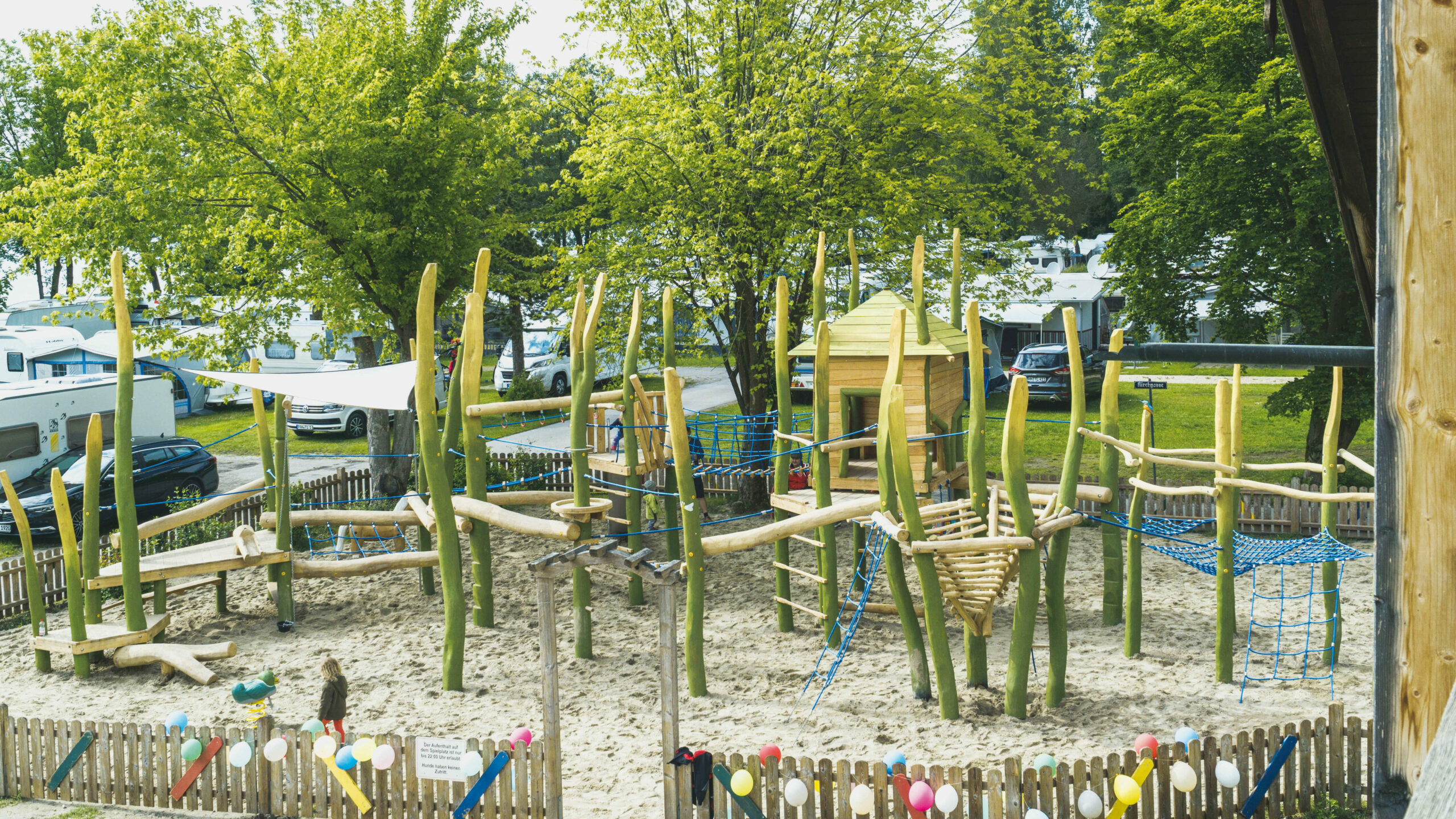 Ein großes Klettergerüst aus Holz und mit vielen Seilen und Netzen in einem Sandkasten, der mit einem kleinen Zaun eingezäunt ist