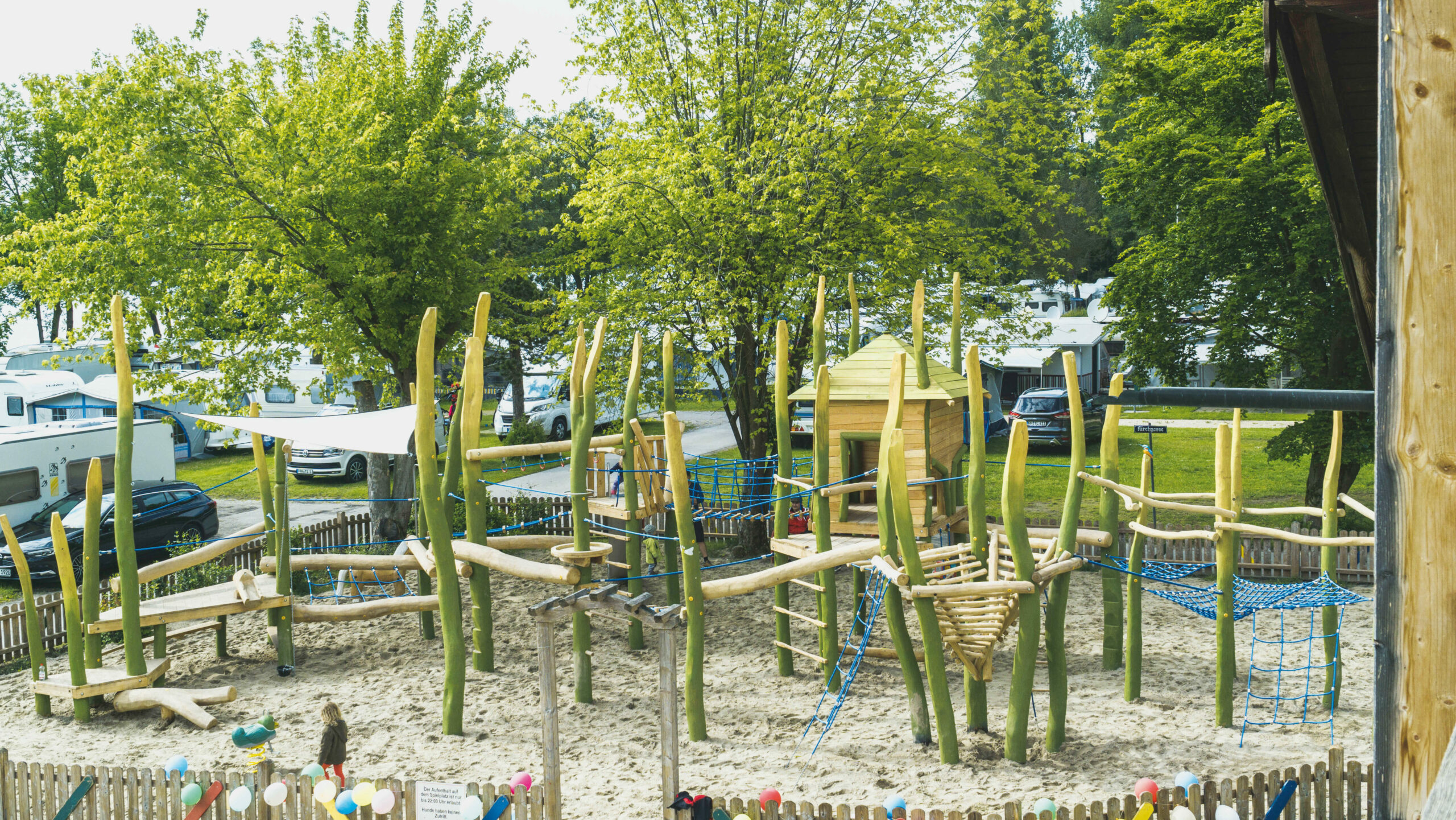 Ein großes Klettergerüst aus Holz und mit vielen Seilen und Netzen in einem Sandkasten, der mit einem kleinen Zaun eingezäunt ist