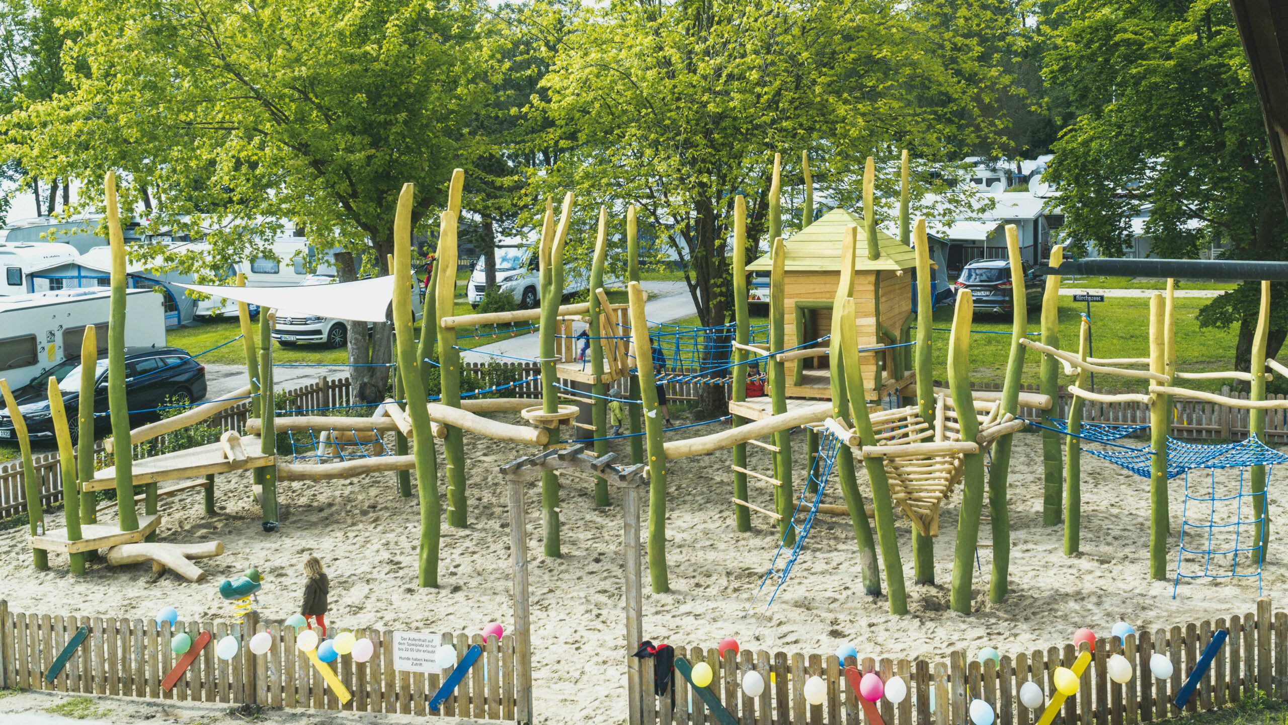 Ein großes Klettergerüst aus Holz und mit vielen Seilen und Netzen in einem Sandkasten, der mit einem kleinen Zaun eingezäunt ist