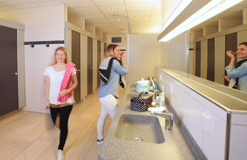 Zwei Frauen in den Sanitäranlagen des Campingplatzes. Links eine Reihe an Toiletten, rechts eine Reihe von Waschbecken mit einem langen, beleuchteten Spiegel.