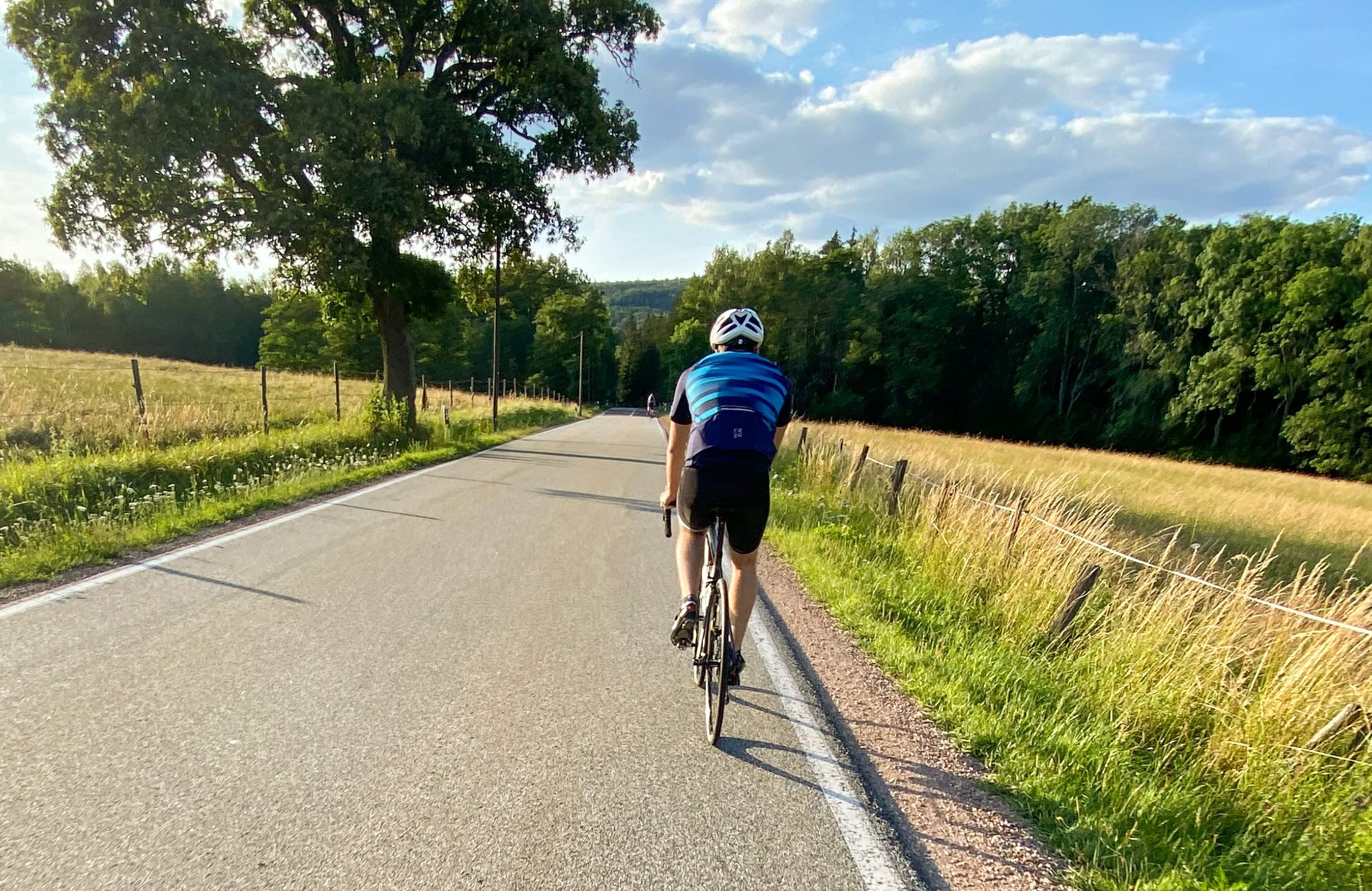 Zwei Rennradfahrer mit Helm und Sportkleidung fährt auf einer sonnigen Straße zwischen zwei Feldern