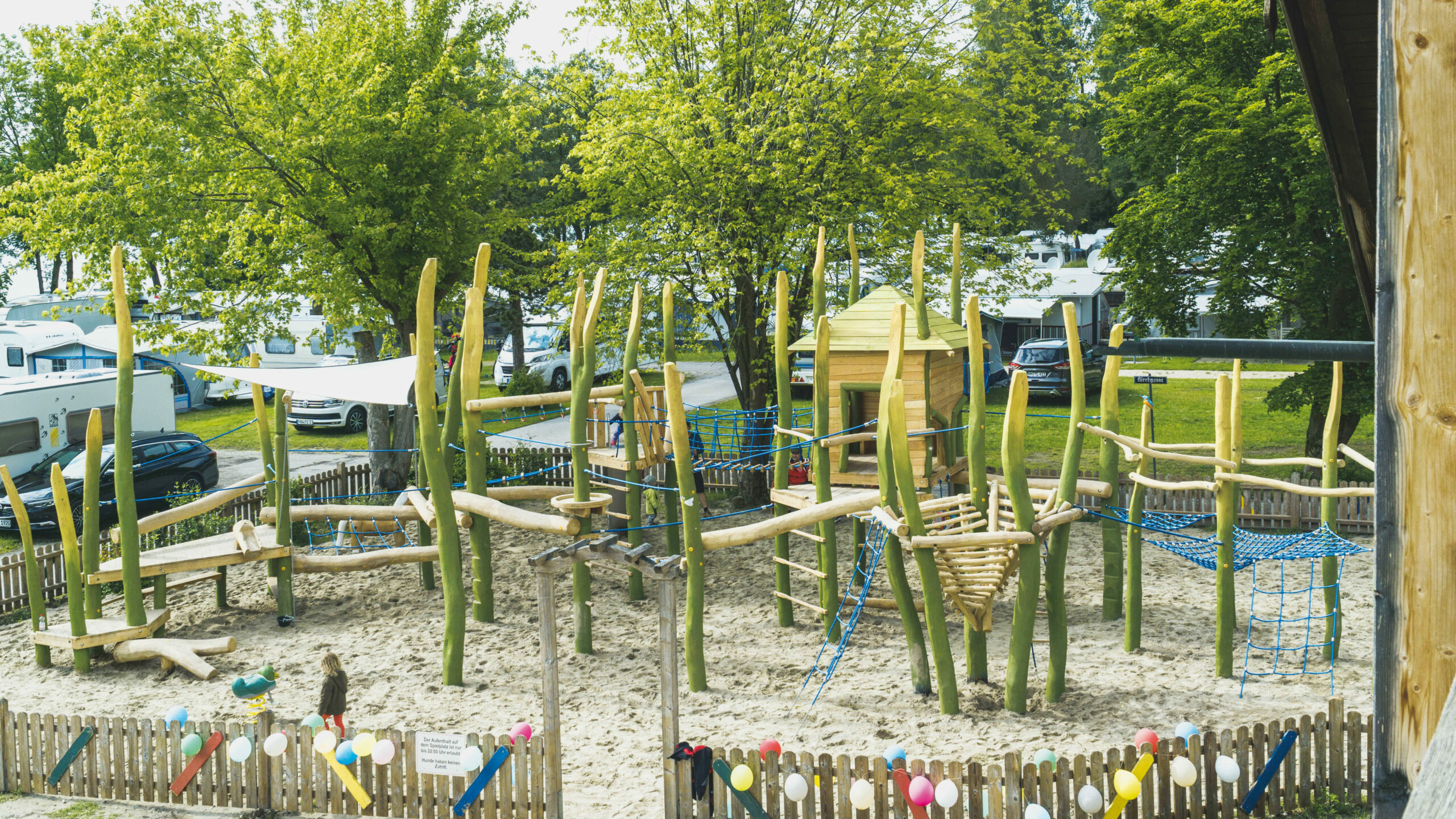 Ein großes Klettergerüst aus Holz und mit vielen Seilen und Netzen in einem Sandkasten, der mit einem kleinen Zaun eingezäunt ist