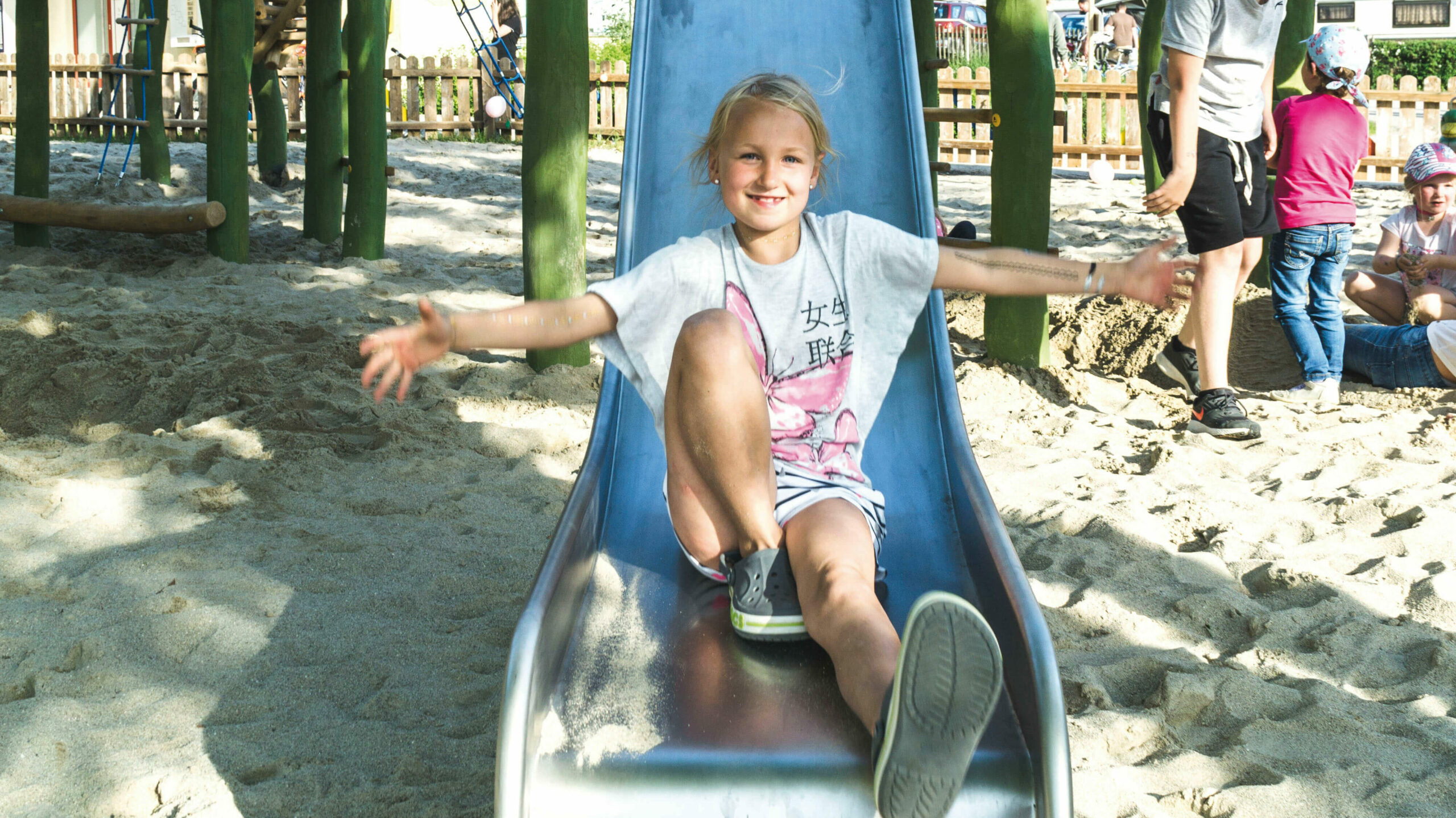 Ein Kind rutscht lachend und mit weit ausgestreckten Armen eine Rutsche auf einem Spielgarten mit Sandboden runter, während mehrere andere Kinder auf einem großen bunten Klettergerüst spielen
