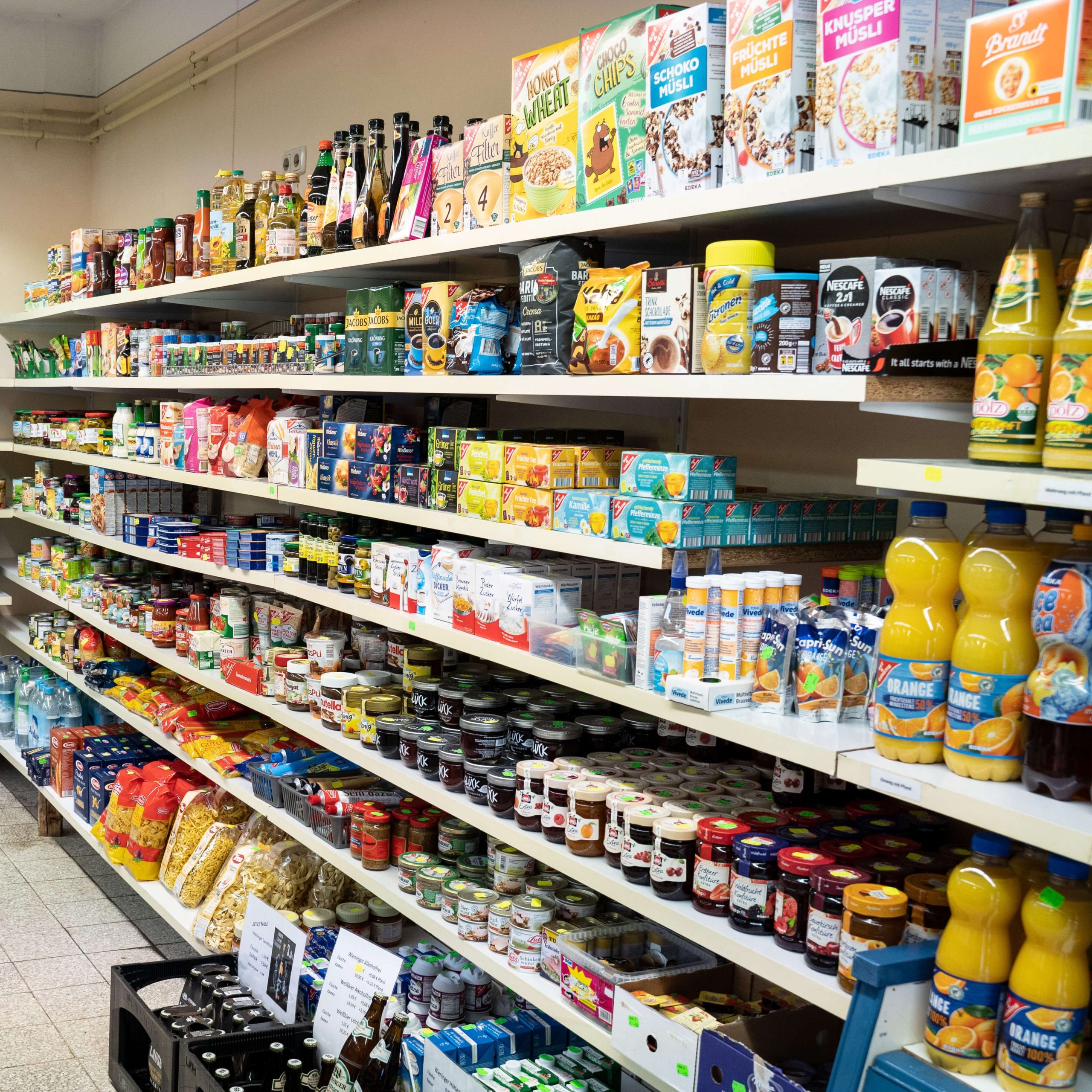 Mehrere voll befüllte Regale eines Supermarkts, links ein Kühlschrank mit Getränken